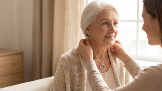 Adult daughter gently fastening a sterling silver generations necklace with interlocking circles around her older mother’s neck in a warm, sunlit room.