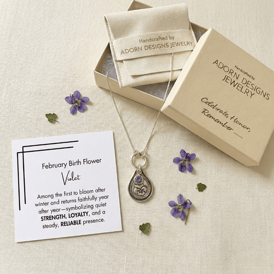 Silver teardrop necklace with pendant, 'February Birth Flower' card, and Adorn Designs jewelry box on a textured beige surface.